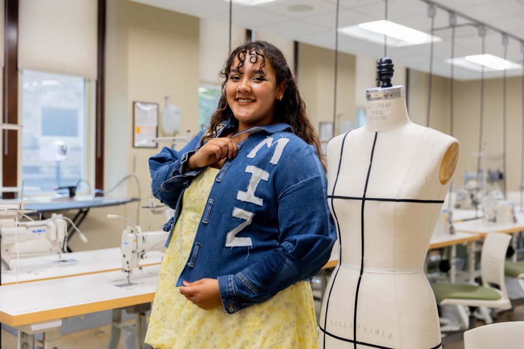 A young woman in a denim jacket with "MIZ" on it poses in a sewing studio, smiling next to a dress form.